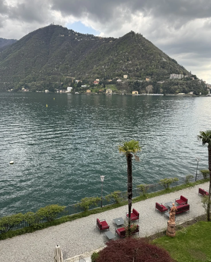A view of Lake Como and its walkways with lounge chairs below. A large mountain stands on the opposite side of the lake.