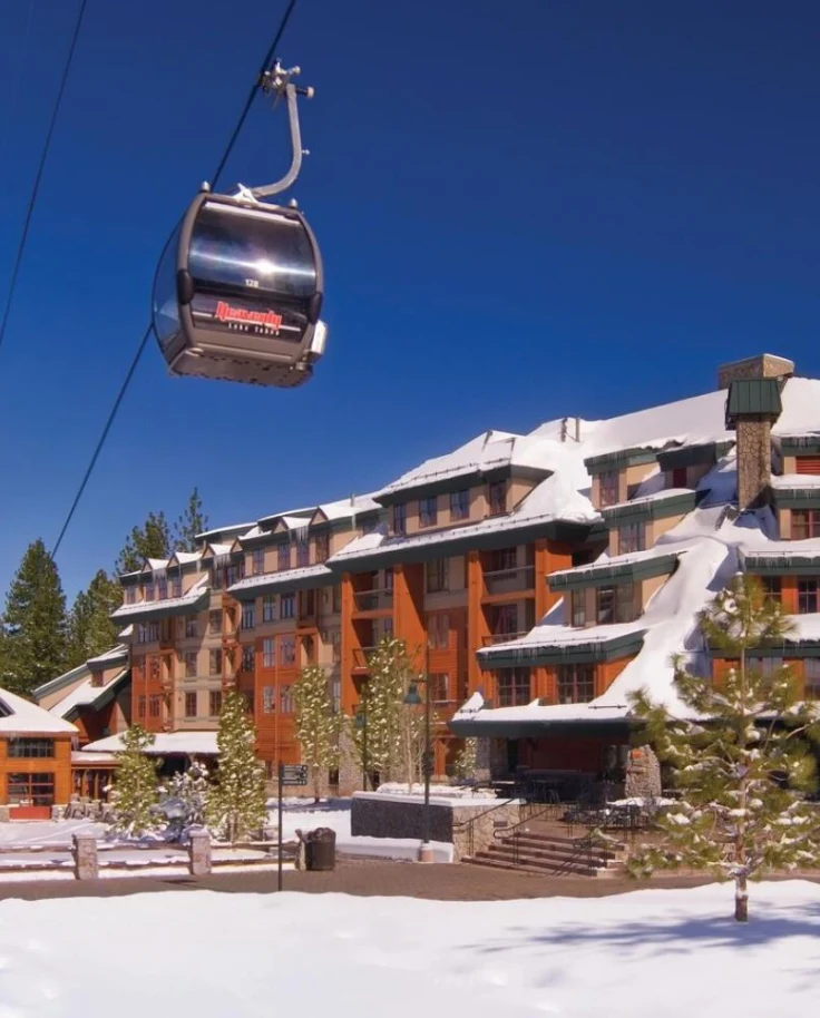 A gondola next a large ski lodge covered in snow and surrounded by pine trees under a clear blue sky.
