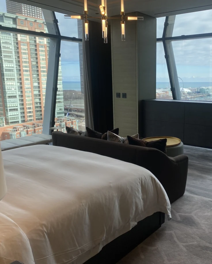 A view of a hotel bedroom with geometric windows, a white bed, modern chandelier lighting, dark wooden credenza and a white chair on the right side.