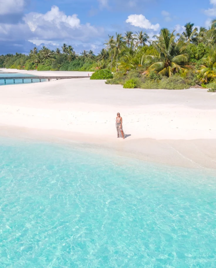 A person stands on a white sandy beach with bright blue ocean water in front and a tropical forest in the background.
