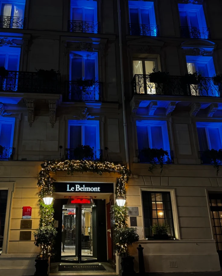 A street view of Le Belmont, with a doorway surrounded by greenery and several floors of blue-lit windows
