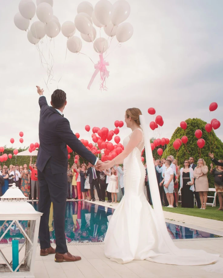 A bride and groom standing next to a pool with wedding guests surrounding and balloons in the air.