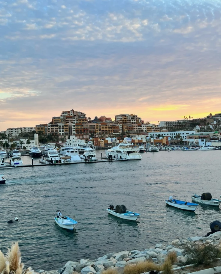 A beautiful harbor with various boats docked and anchored around a mountainous and coastal town, with a yellow sunset and clouds in the distance.