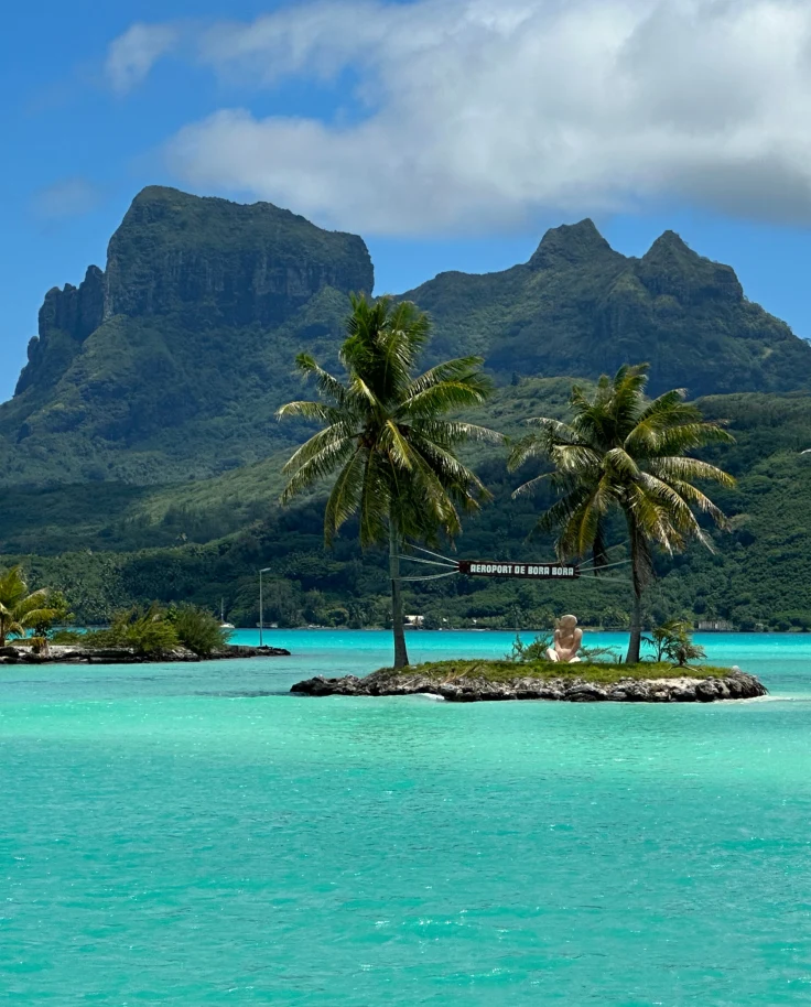 A view of a bright teal blue ocean with a small island with two palm trees and a large mountain in the distance on a partly cloudy day.