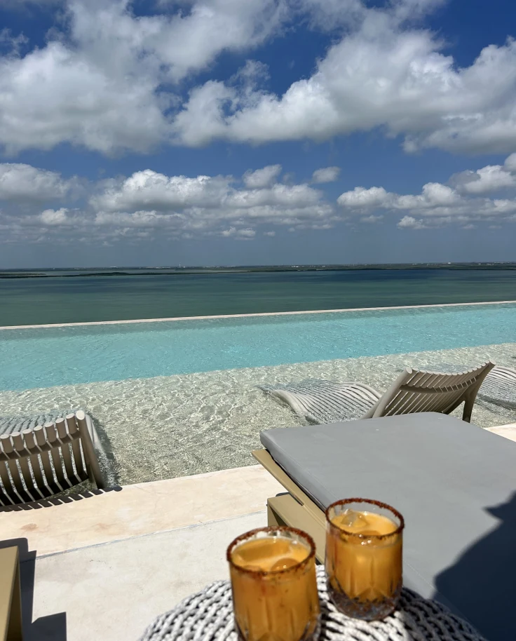 A hotel balcony and infinity pool scene with lounge chairs overlooking the sea and sky on a sunny day.