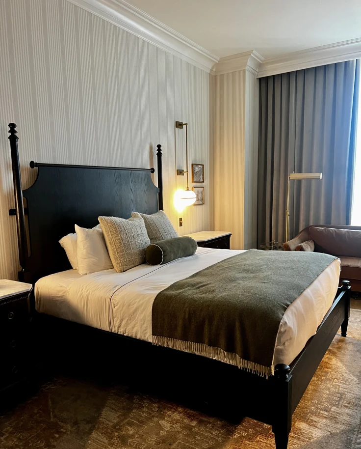 A view inside a hotel room with white pinstriped walls, a dark wood bedframe and headboard with white bedding and dark curtains.