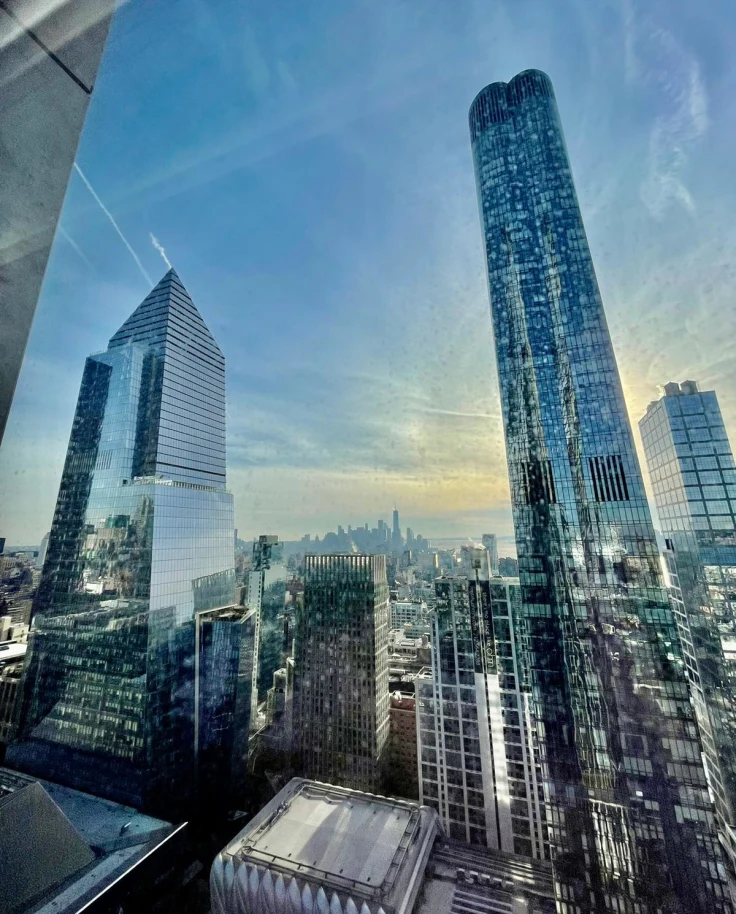 A view of the reflective skyscrapers in New York City with the sun peaking out of the clouds in the distance.