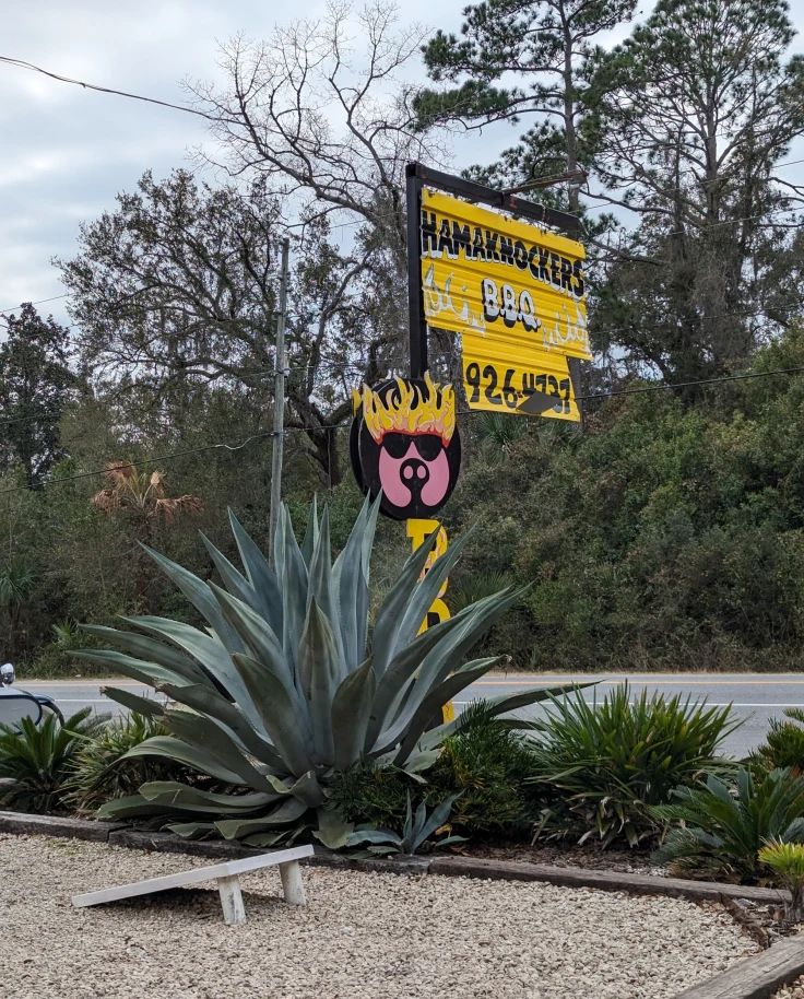 A yellow sign for a restaurant surrounded by trees and aloe vera plants on the side of the road outside.