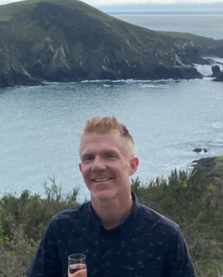 A person standing by a railing smiling for a photograph with a view of the water and a mountain in the distance