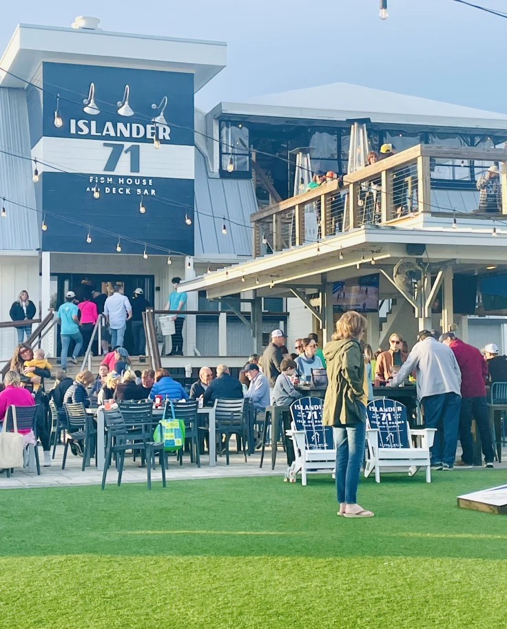 A group of people dining and socializing outside on a patio and lawn. There are string lights, a restaurant exterior and a wooden balcony in the surrounding areas.
