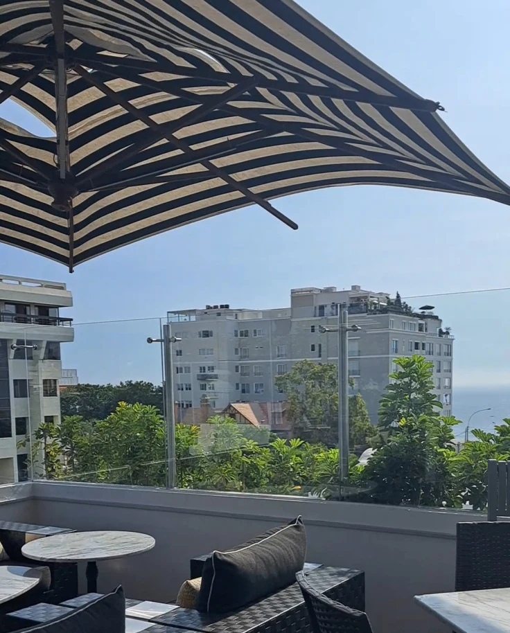 A view of the rooftop with black chairs and cabana seating underneath a black and cream striped umbrella. There are buildings and trees in the distance.