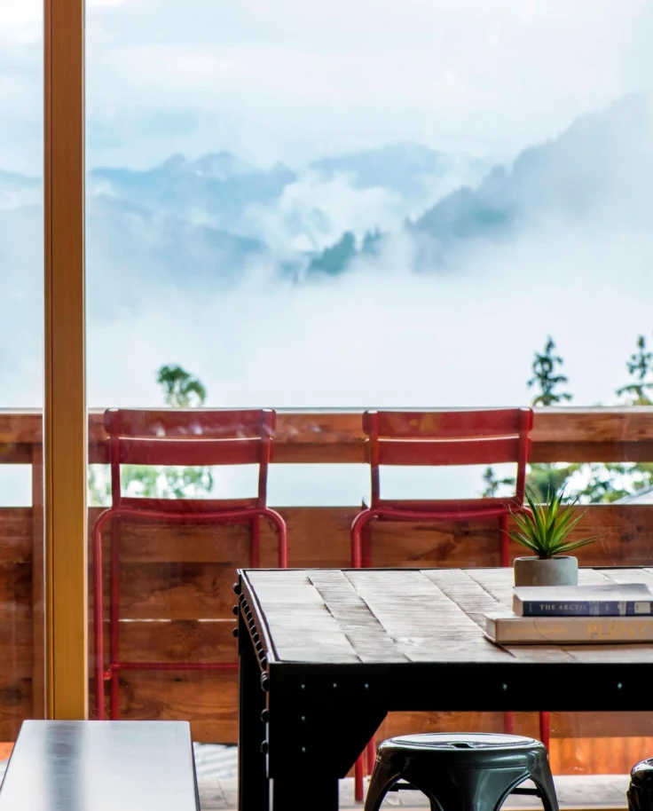 A black table and red stools against a wooden ledge with mountains and clouds in the background.
