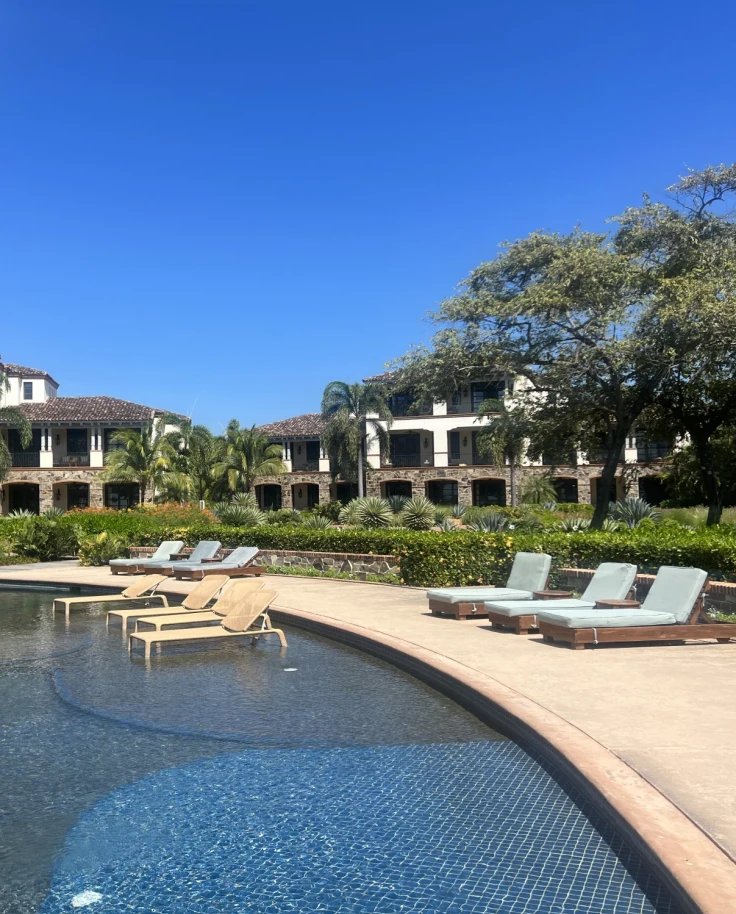 Pool area at a resort with sun loungers around the pool and some sun loungers in the pool, with green landscaping and two-storey buildings behind
