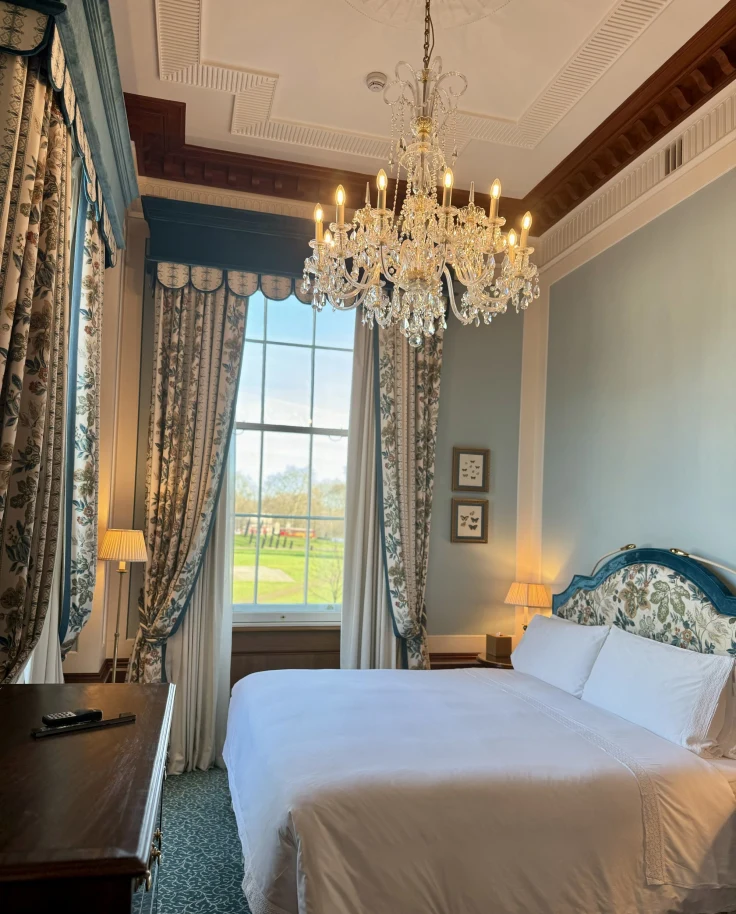 A picture of a hotel suite, complete with a white bed, blue-toned and patterned headboard, crystal chandelier, wooden table, lamps that are turned on, patterned curtains and blue walls.