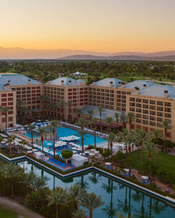 An aerial view of a resort surrounded by palm trees, a pool with lounge chairs and umbrellas, a bar a body of water and mountains beneath a golden sunset in the distance.