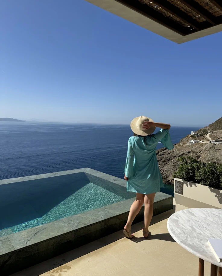Eliza in a blue dress and sun hat standing by an infinity pool overlooking the ocean.