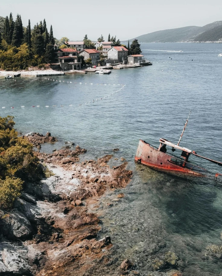 Shipwreck Beach in Rose at the entrance to the Bay of Kotor, with a red boat partially submerged in water off of a tree-covered hillside.