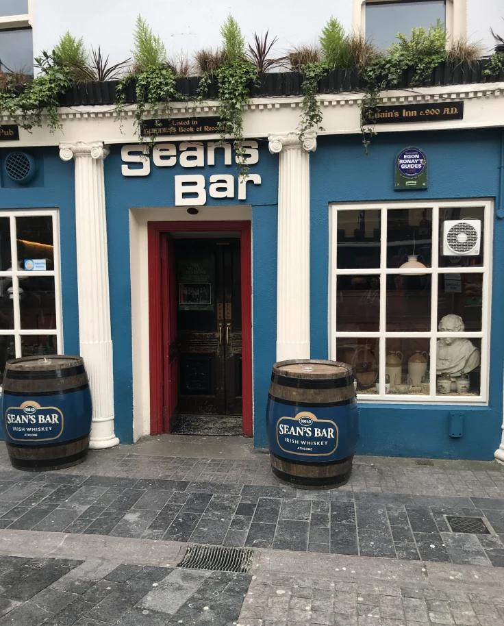 An image of the exterior of Seans Bar in Ireland with a blue and white facade and two barrels on either side of the entrance.