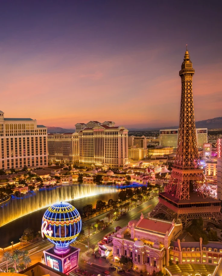 An aerial view from the Encore Tower Suites, highlighting the Bellagio Hotel and fountain, mock Eiffel Tower and other Las Vegas buildings, some lit up before a dusk sky.