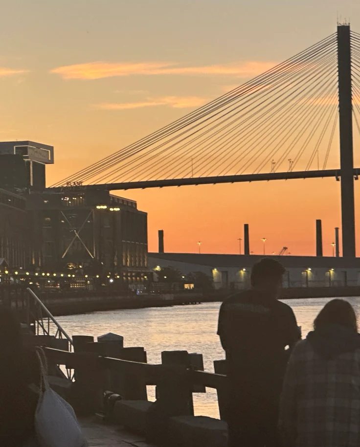 Savannah Riverfront Walk at sunset, with people walking along the water and a bridge in the background - Cara Clinton-Vollmer