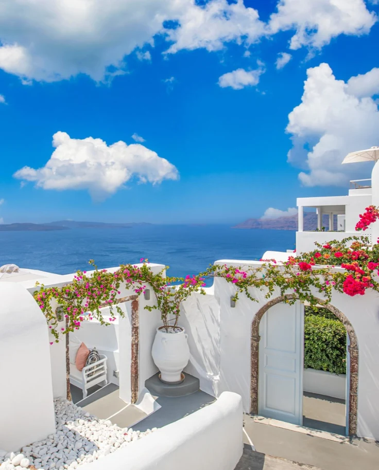 A picture of a white terrace with pink and green flowers, a white pot, an archway and a cloudy blue sky above with the sea in the background.