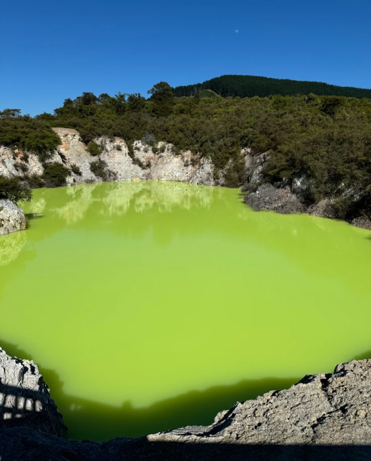 A vibrant green geothermal pool is surrounded by rocky terrain under a clear blue sky.