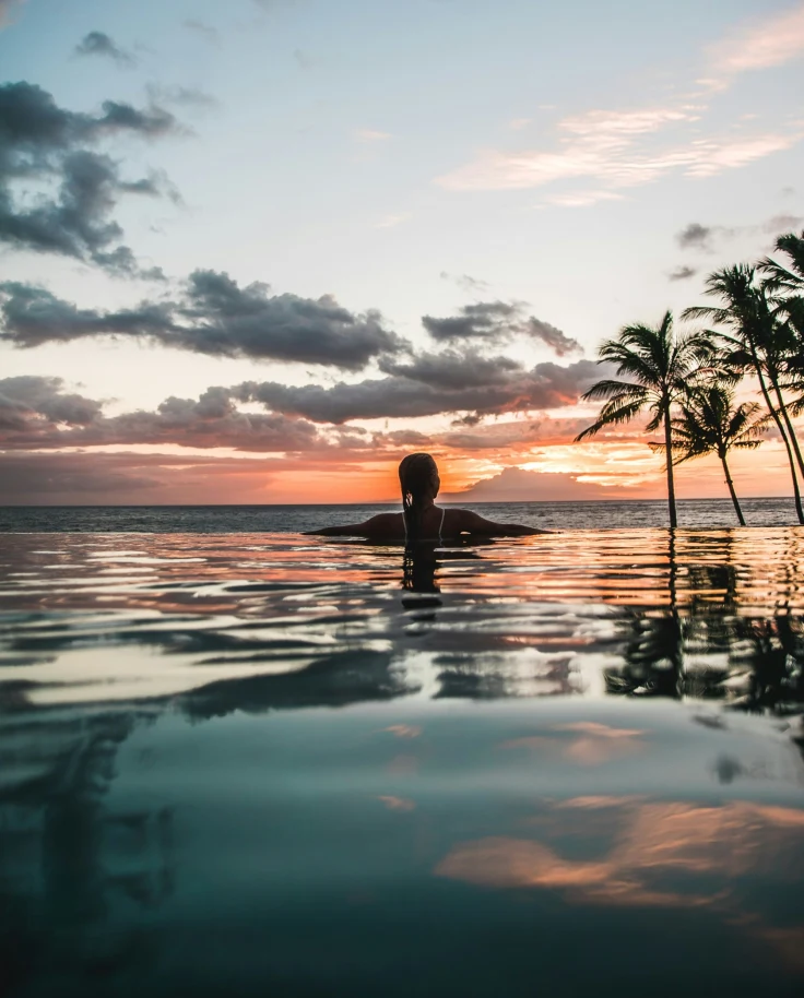 A serene sunset scene with a person immersed in water, silhouetted against a sky ablaze with vibrant sunset hues and palm trees.