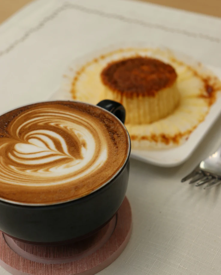 Coffee latte art (wing heart) and cheesecake on a plate sitting on top of a table.