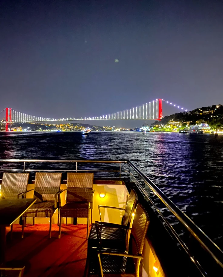 A beautiful view of chairs on a balcony overlooking a river and colorfully arched bridge in the distance at nighttime.