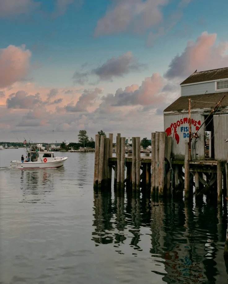 The image shows a pier with a white building with a red painted sign saying 'Gosmans', at sunset with pink fluffy clouds.