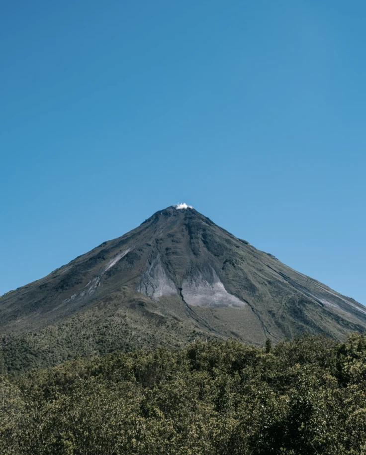 A large volcano surrounded by trees and blue sky.