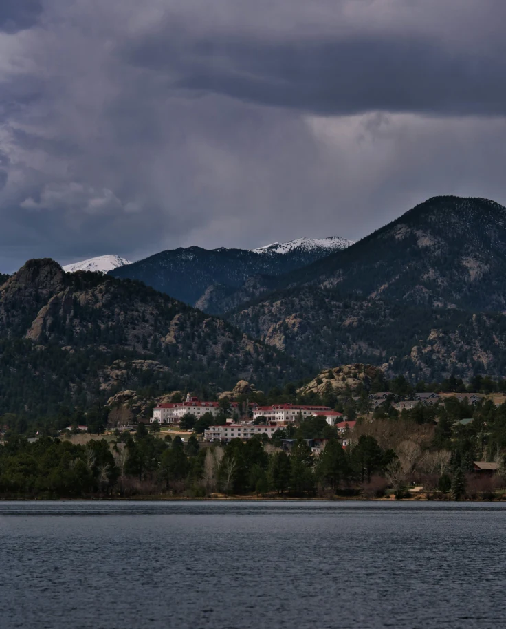 A view of a dark body of water with a white villa nestled into a green mountain under a cloudy grey sky.
