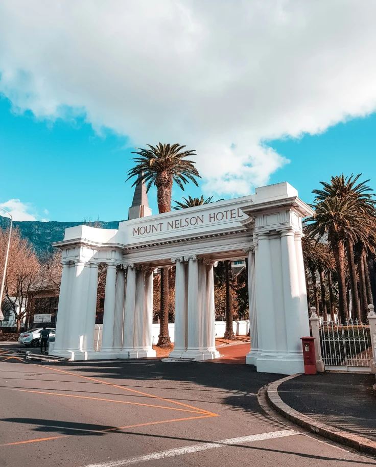 A beautiful entrance with white arches to the Mount Nelson Hotel, surrounded by palm trees and a road.