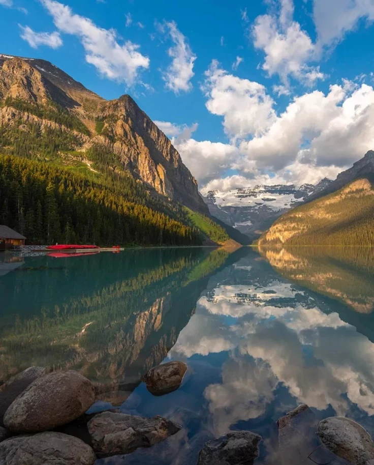 The image shows a serene lake reflecting mountains and clouds, with a cabin and canoes to the left, set in a tranquil forested area.