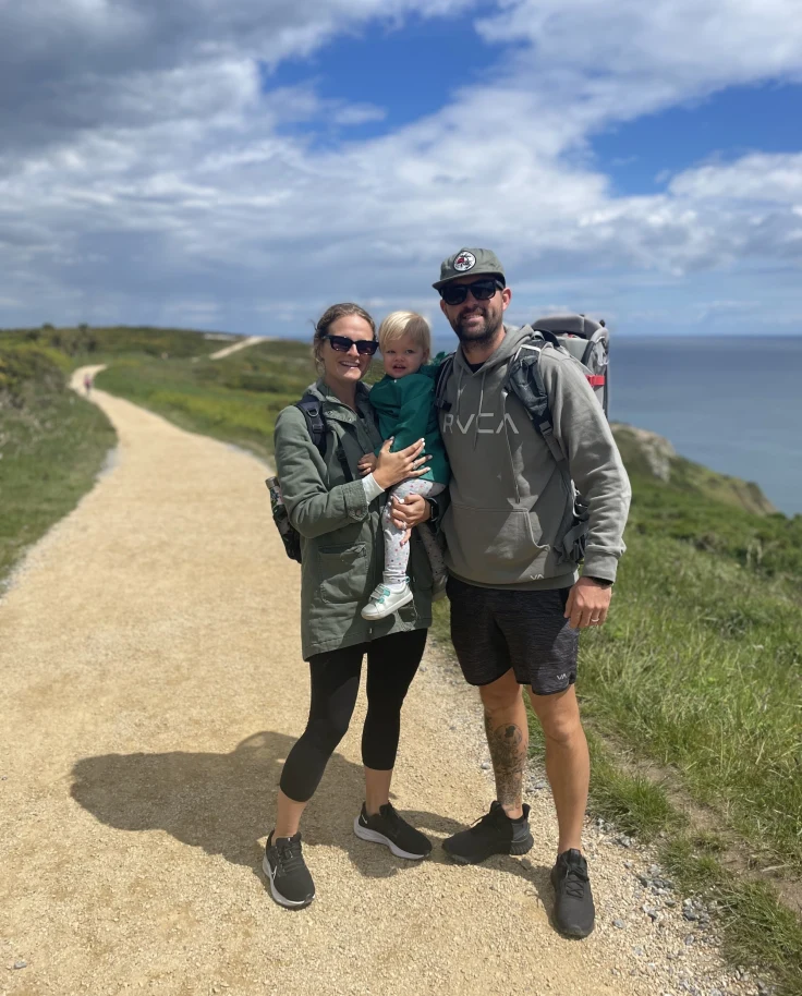 The image shows two adults and a child standing on a dirt path with a scenic ocean view in the background.