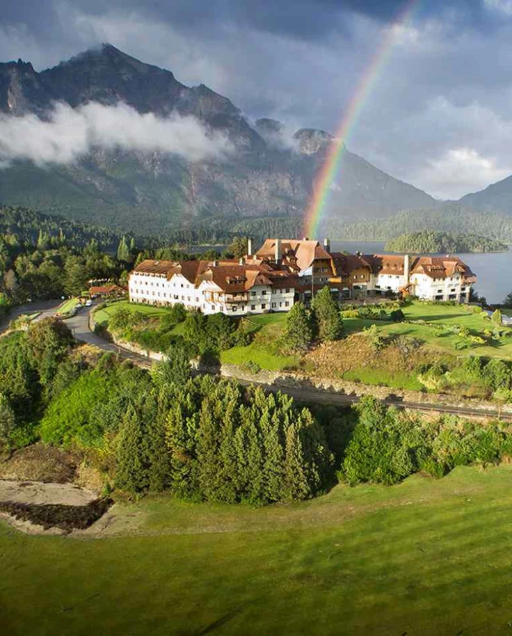 Beautiful scenery with a large estate on a hill by a lake and mountains with a rainbow overhead.