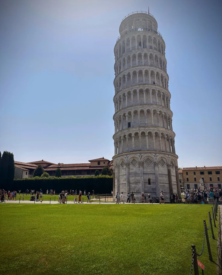 A view of the leaning tower of Pisa during the day time. There is a manicured, green lawn in the front and a clear blue sky in the background.