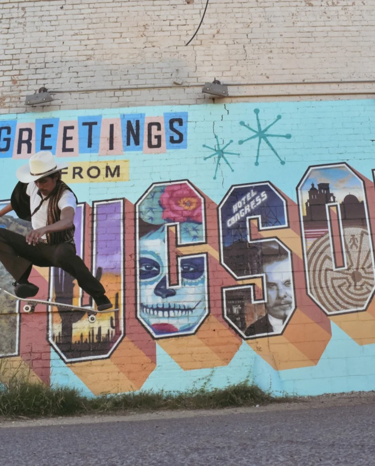 Man in a cowby hat on a skateboard midair, in front of a mural saying Greetings from Tucson.