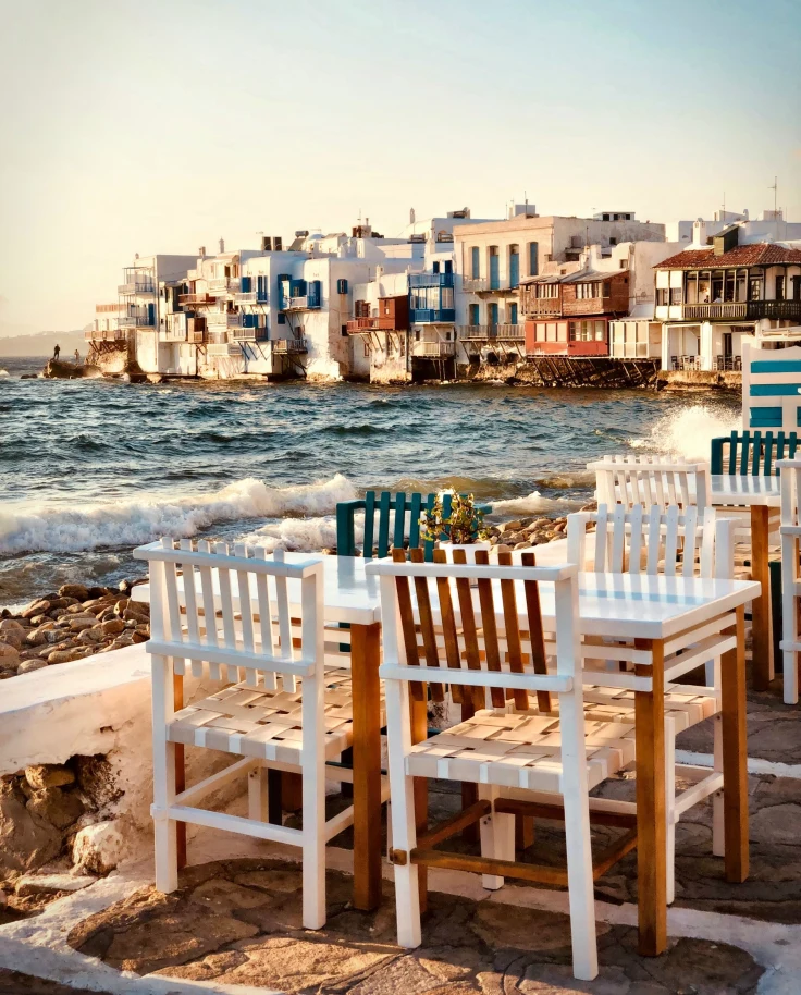 Tables and chairs near the blue water with Greek architecture in the background.