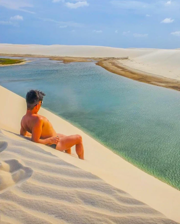 The image depicts a person enjoying a serene view of a calm water body from the edge of a white sand dune.