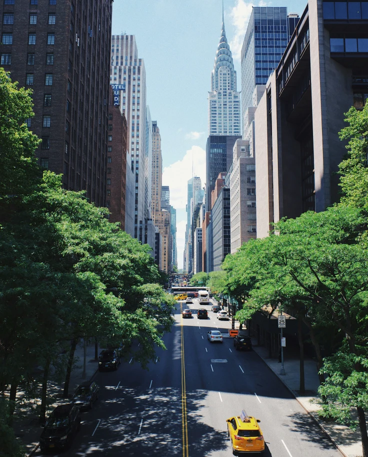A bustling New York City street with greenery and the Chrysler Building in the distance under a clear blue sky.