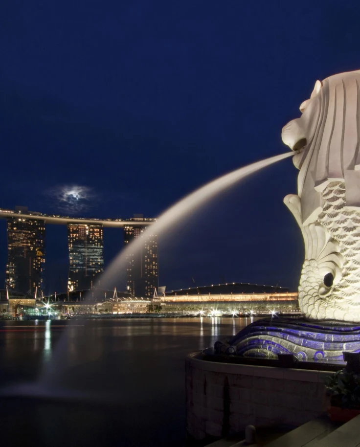 An image of a large white sculpture with water spraying from the mouth into the ocean and three tall skyscrapers illuminated in the background.