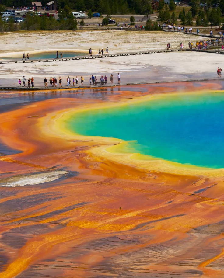 An orange, yellow, green and blue geysir in Yellowstone National Park with people standing around the outskirts.