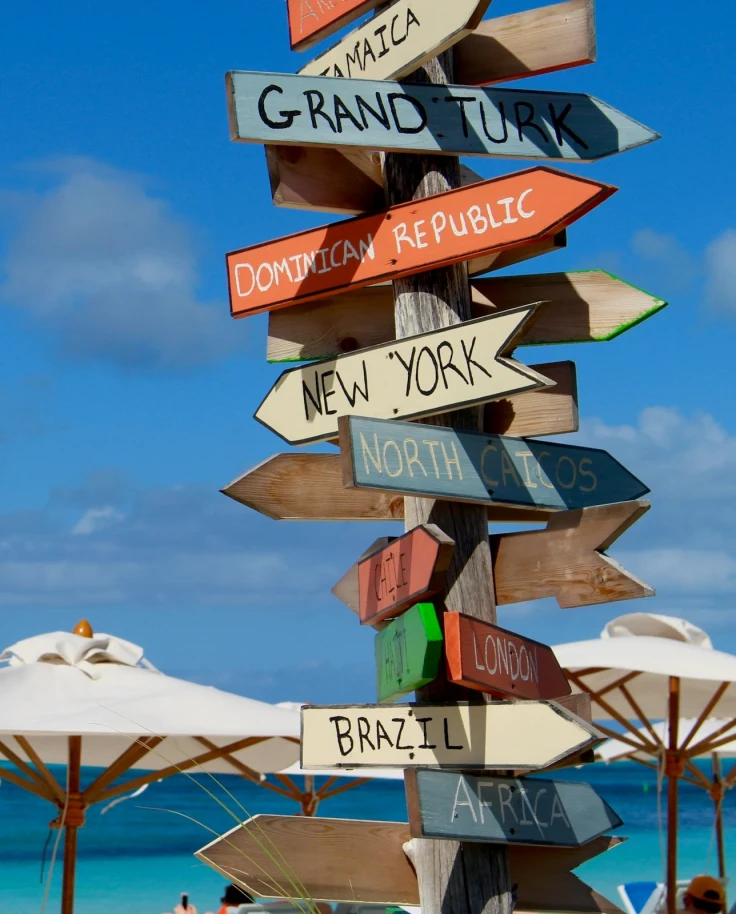 Signages pointing in different directions towards major cities and white patio umbrellas in the seashore of Providenciales.