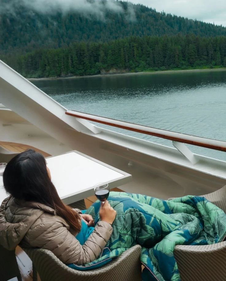 A person sitting on a deck chair on a cruise ship, wrapped in a blanket, looking out at a calm river and forested hills with misty clouds above.