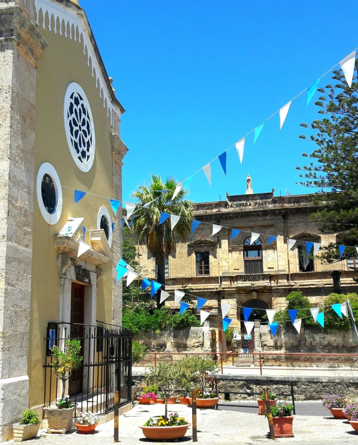 A concrete walkway lined with plants leading to a yellow building, with blue and yellow flags flying outside, which you might see on a Sicily itinerary, 10 days. - Ali Lefebvre
