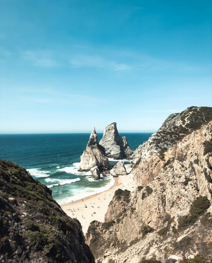 The image displays a coastal landscape with majestic rock formations on a sandy beach under a clear blue sky.