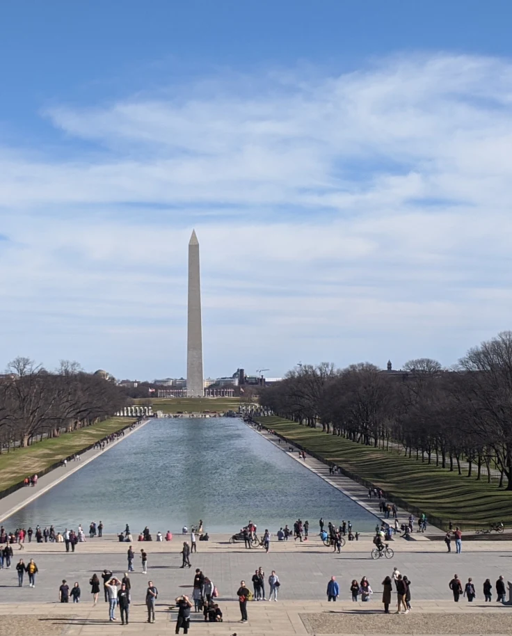 A view of a long body of water with green spaces and people with a tall monument in view in Washington D.C.