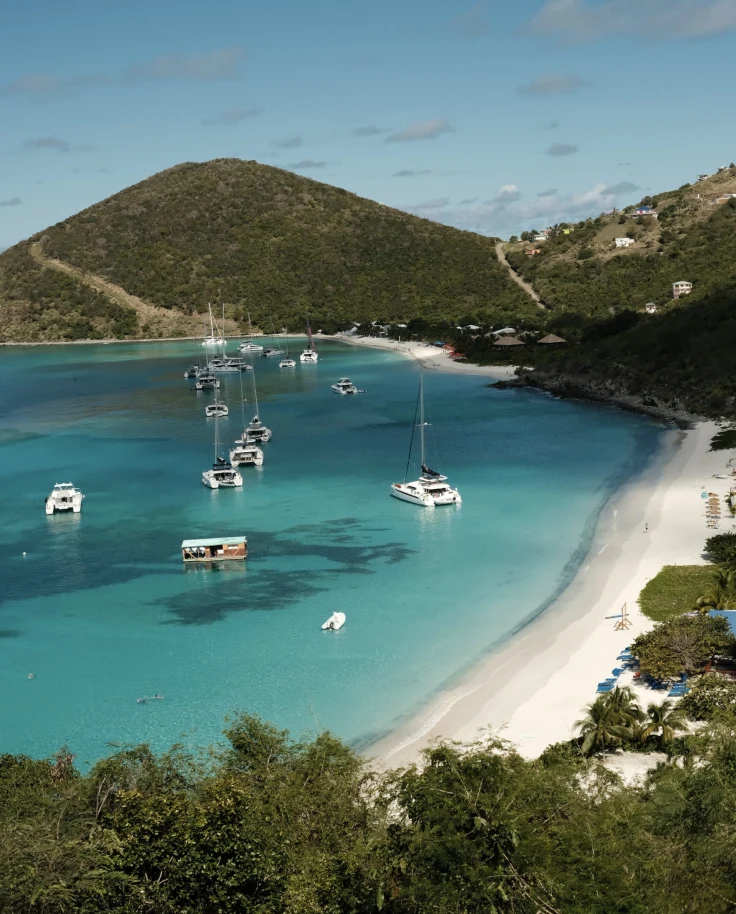 A scenic coastal landscape with boats anchored in clear blue waters near a curved sandy beach, surrounded by lush green hills.