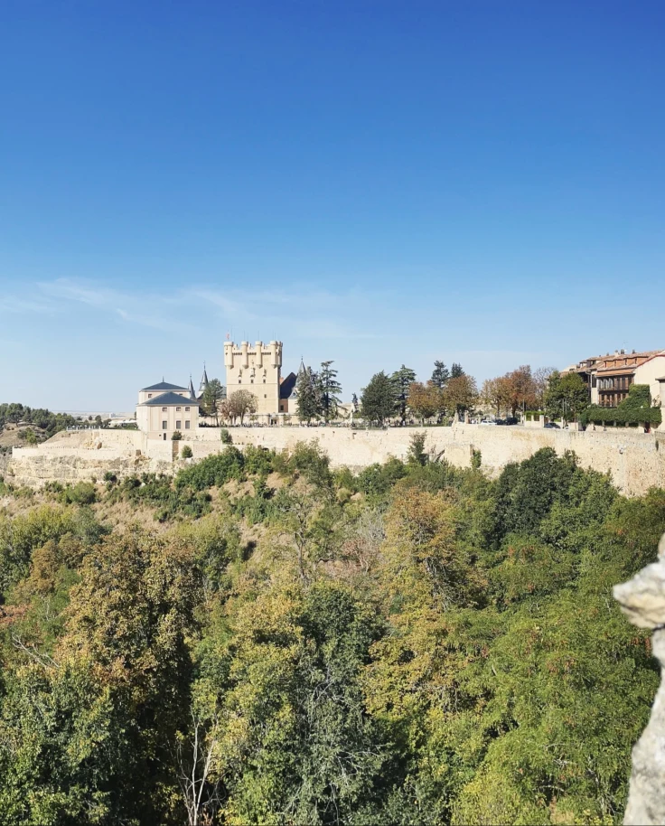 A historic stone building with towers is surrounded by lush greenery under a clear blue sky.